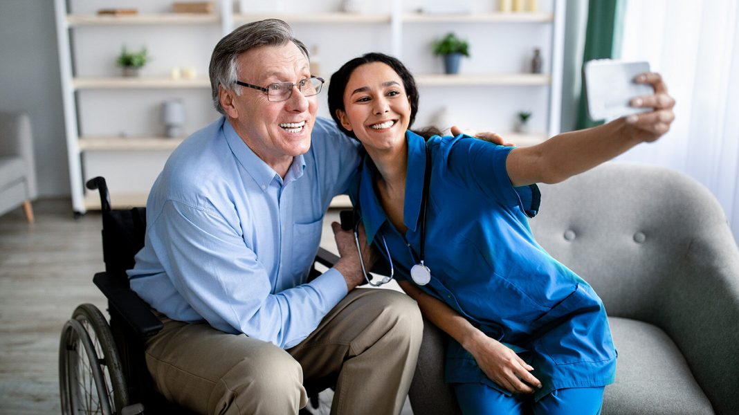 Cheerful impaired senior man in wheelchair taking selfie with young female doctor at retirement home. Disabled older patient making photo with nurse, having fun together indoors